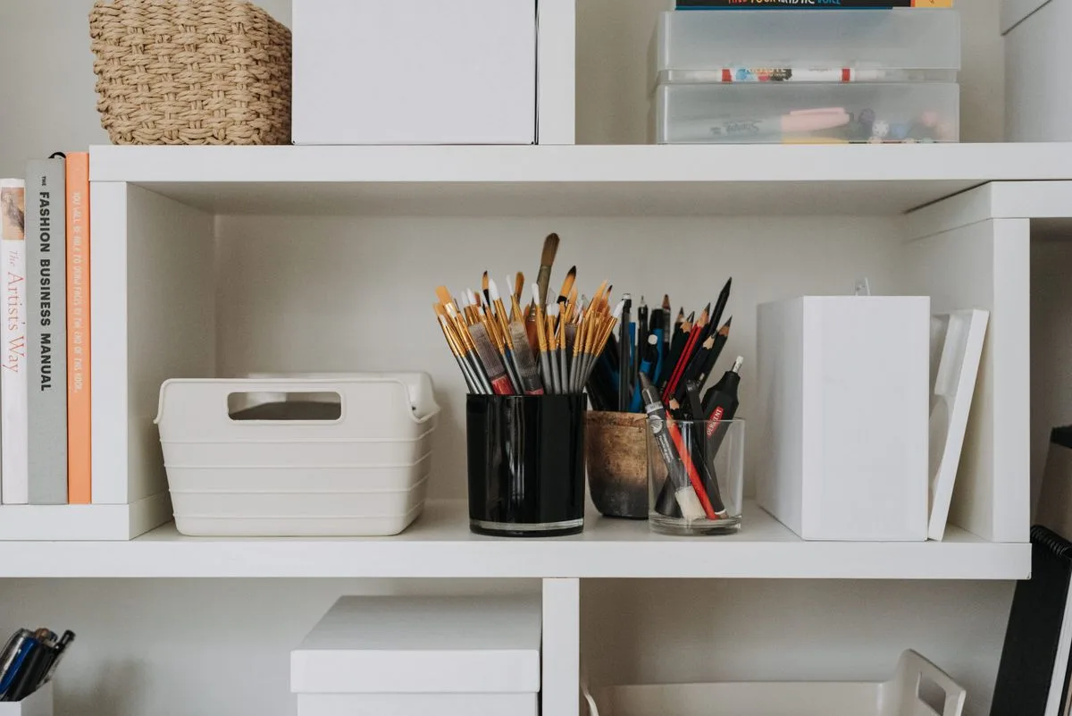 Paintbrushes, pencils and art materials organised on white shelves.