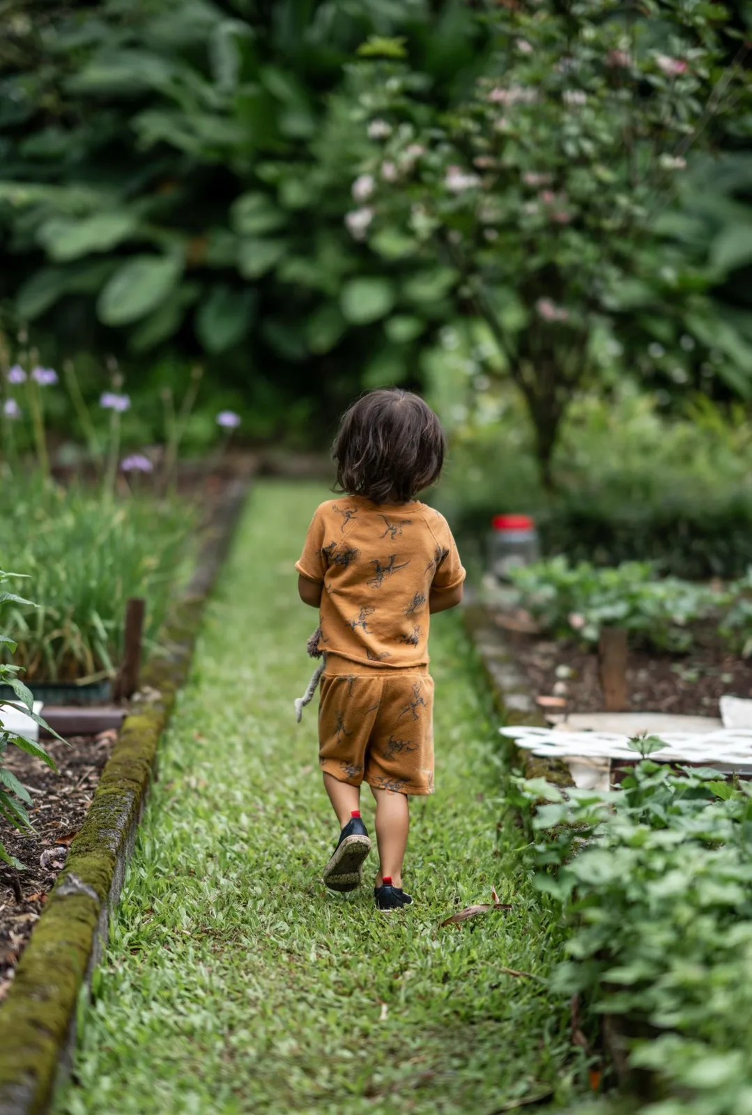 A young child walks along a garden path, surrounded by lush greenery and foliage.