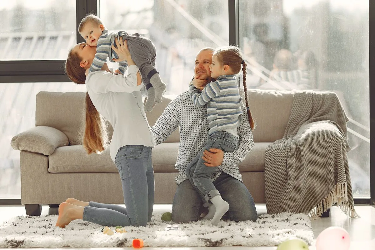 Cheerful mother kissing little boy while father hugging adorable daughter when family sitting on floor in rainy day against window