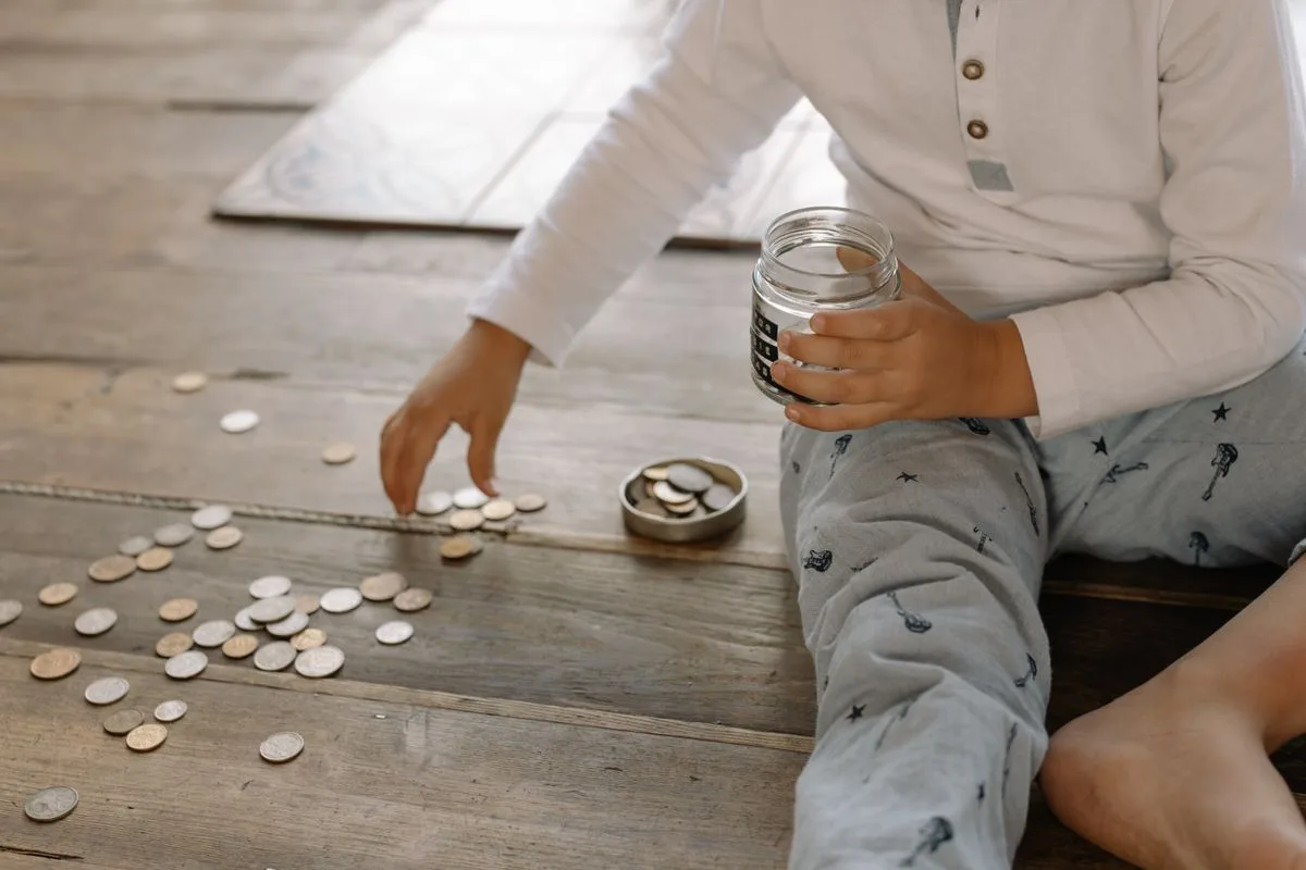 A young child collects coins into a jar on a wooden floor.