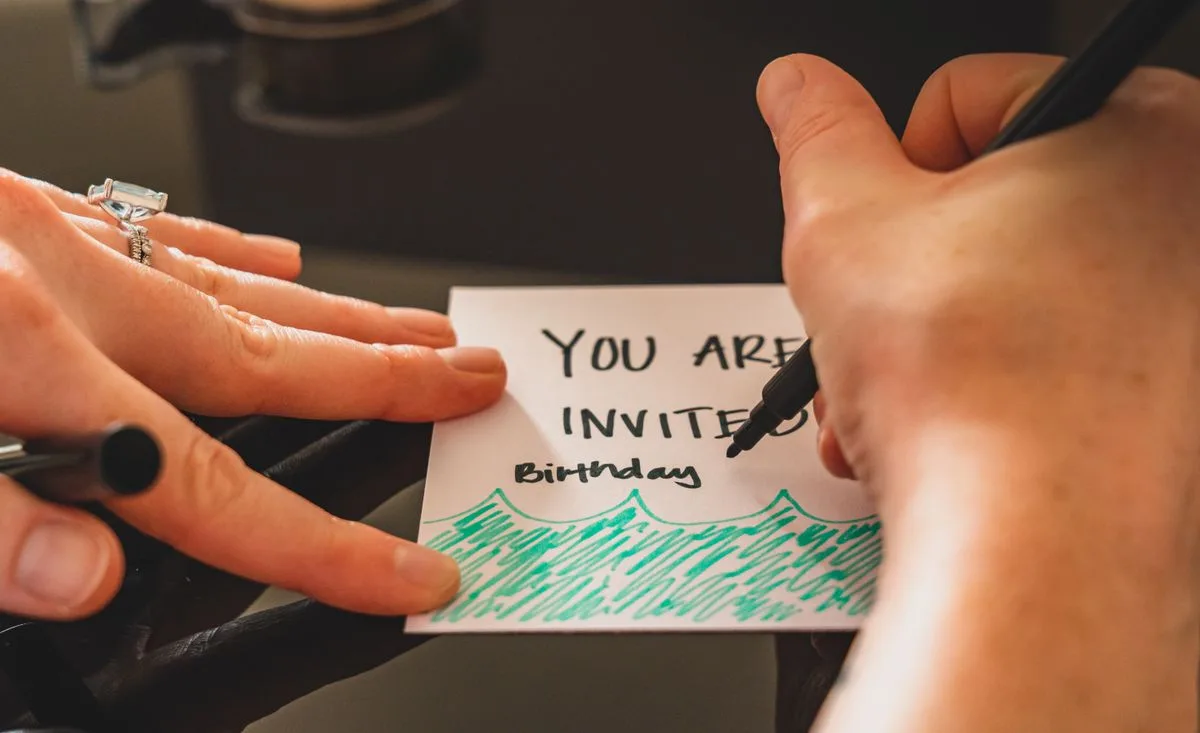 Hands writing a personalized birthday invitation card using a marker on paper.