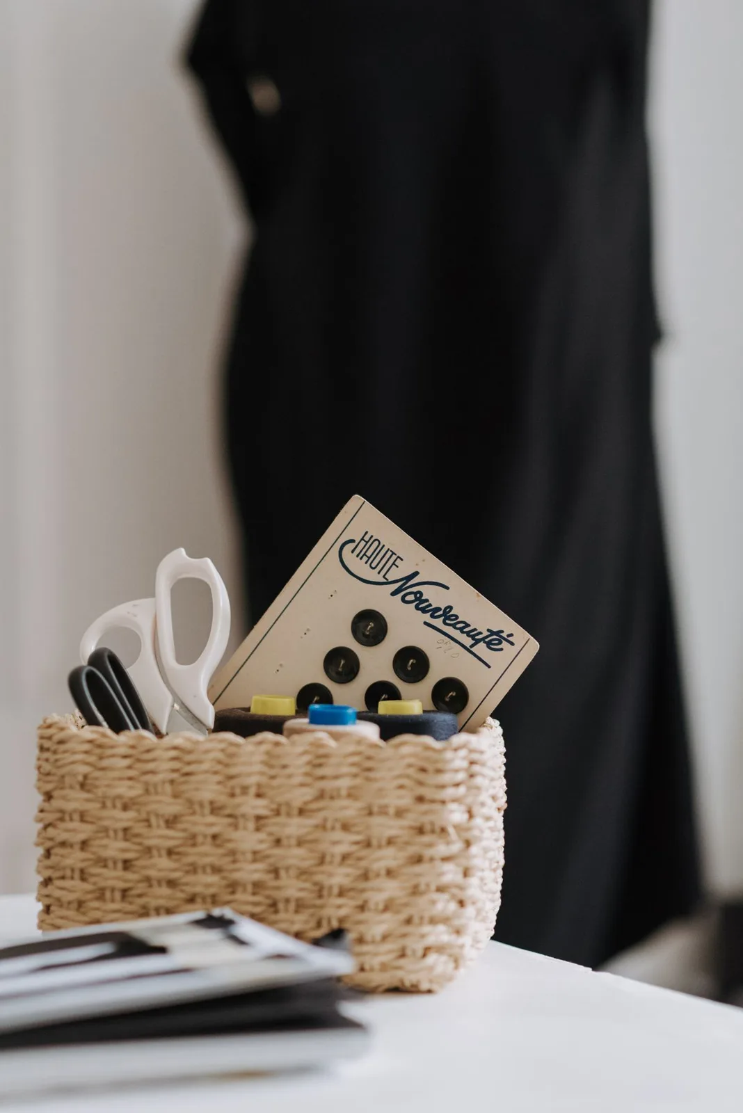 Wicker basket with scissors thread spools and black buttons placed on white desk in light professional workshop with various supplies