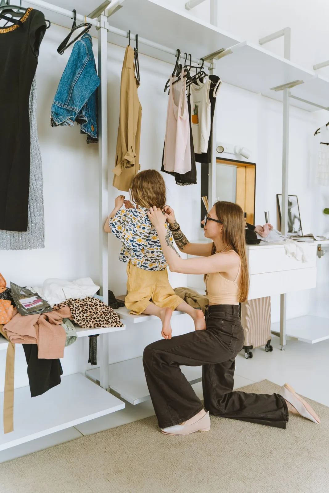 A mother lovingly helps her child dress in a stylish, modern wardrobe setting indoors.