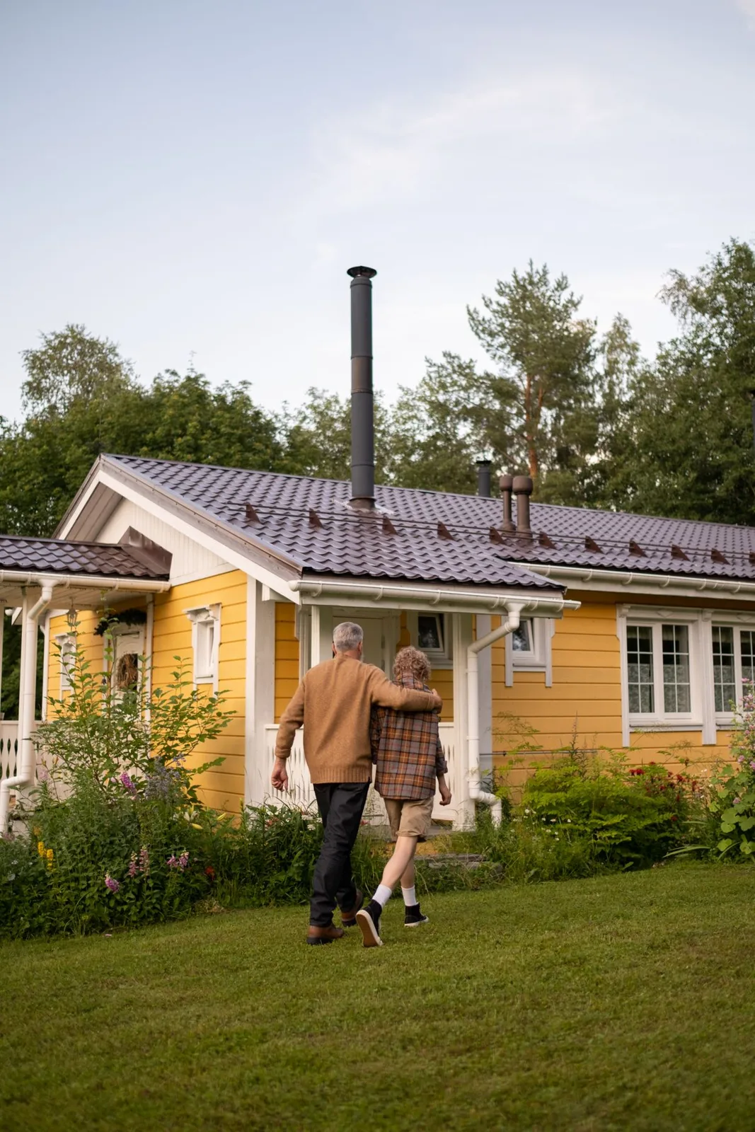 A serene moment of togetherness as a grandparent and child walk by a charming yellow cottage in lush greenery.
