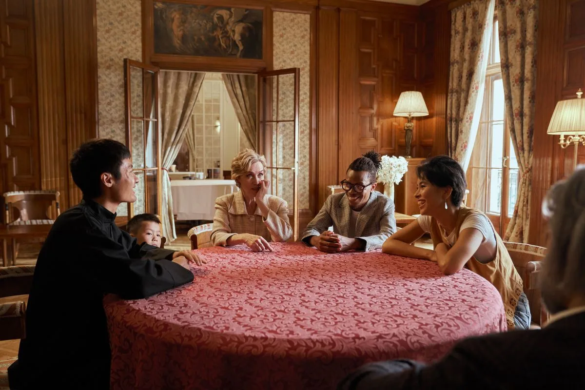 Diverse group of adults and children conversing at a round table in a luxurious mansion setting.