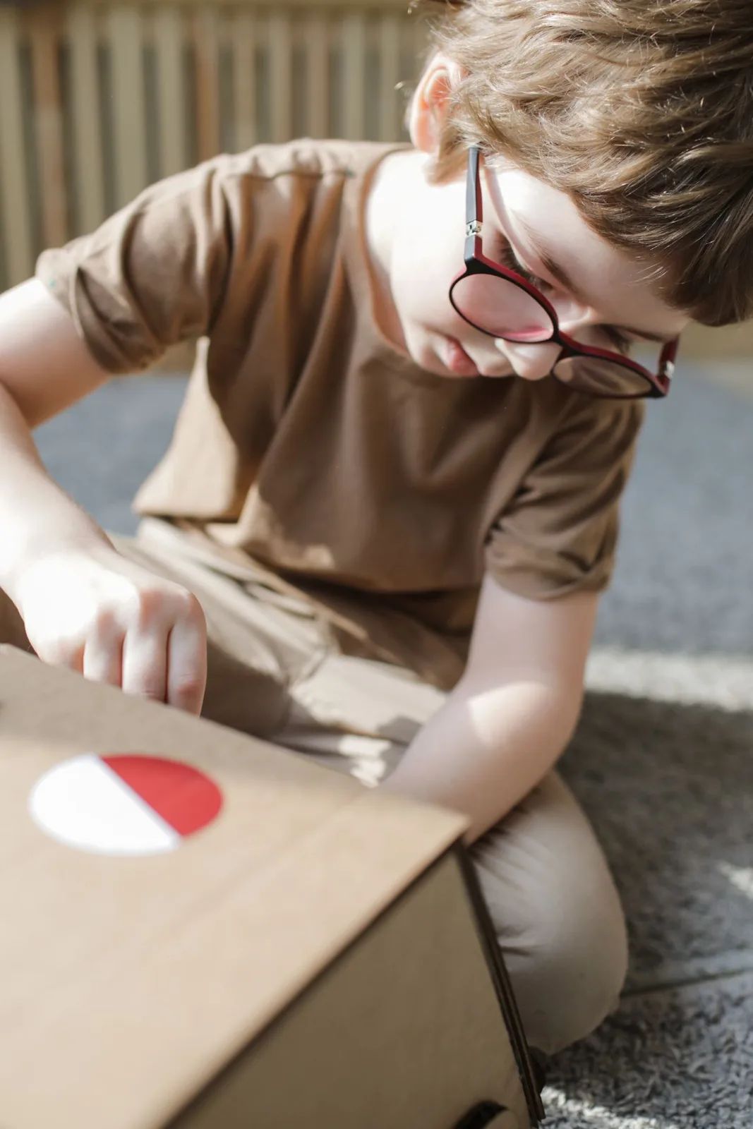 Child in brown shirt and glasses plays with a DIY cardboard box indoors.